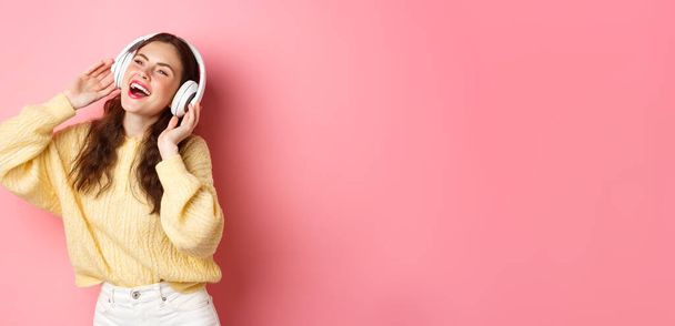 Beautiful modern girl singing favorite song, listening to music in wireless headphones, smiling and dancing, standing over pink background. - 写真・画像