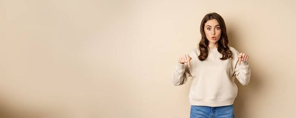 Enthusiastic attractive girl pointing fingers down, showing advertisement, link or promo below, standing over beige background. - Φωτογραφία, εικόνα
