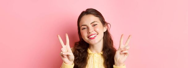 Nahaufnahme einer schönen jungen Frau, die das Peace V-Zeichen zeigt und glücklich in die Kamera lächelt, helles Glam-Make-up trägt und vor rosa Hintergrund steht. - Foto, Bild