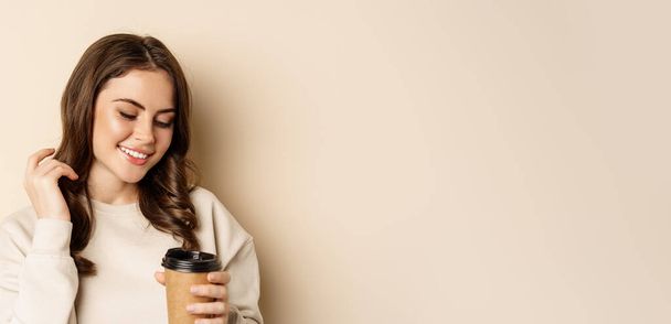 Takeaway and cafe concept. Beautiful feminine woman smiling, holding cup of coffee, posing against beige background. Copy space - Foto, Imagen