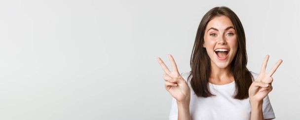 Portrait of beautiful and happy brunette girl smiling and showing peace gestures, standing white background excited. - Photo, image