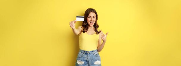 Portret brunetki pokazującej swoją kartę kredytową i uśmiechniętej, ubranej w letnie ubrania, koncepcję banku, pieniędzy i zakupów, stojącej na żółtym tle. - Zdjęcie, obraz