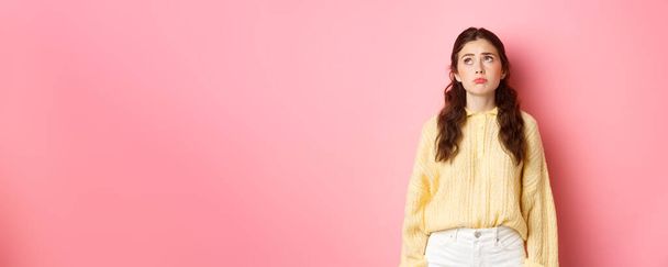 Image of upset and gloomy young woman pulling sad face, looking up with regret or jealousy, standing disappointed against pink background. - Photo, image