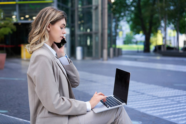Frau telefoniert und arbeitet mit Laptop, sitzt im Stadtzentrum auf der Straße und trägt Anzug. - Foto, Bild