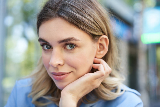 Close up portrait of young woman, tuck hair behind ear, looking flirty and smiling, sitting in blue shirt outdoors on street. - Фото, изображение