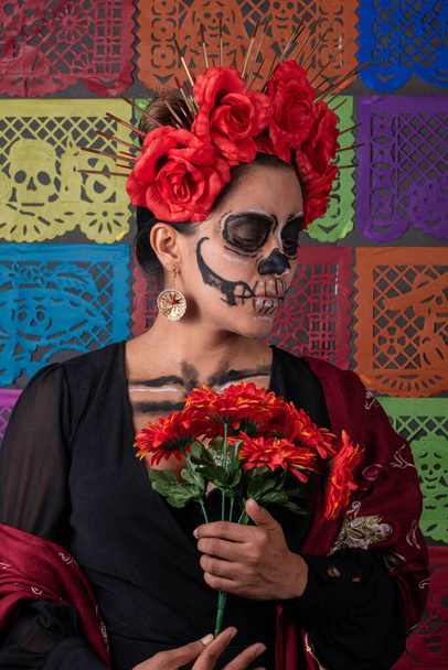 Mexican woman in traditional make up and Catrina costume, holding an orange rose. Colorful cut paper background - Photo, image