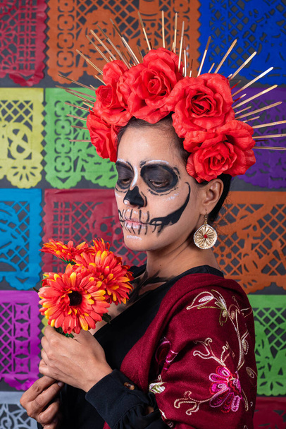 Mexican woman in traditional make up and Catrina costume, holding an orange rose. Colorful cut paper background - Photo, Image