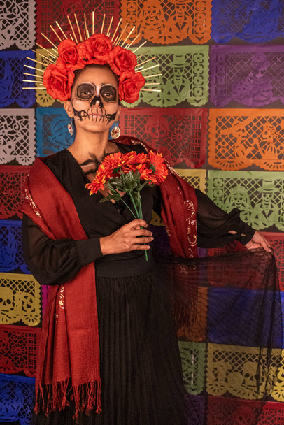 Mexicaanse vrouw in traditionele make-up en Catrina kostuum, met een boeket van oranje bloemen. Kleurrijke gesneden papier achtergrond - Foto, afbeelding
