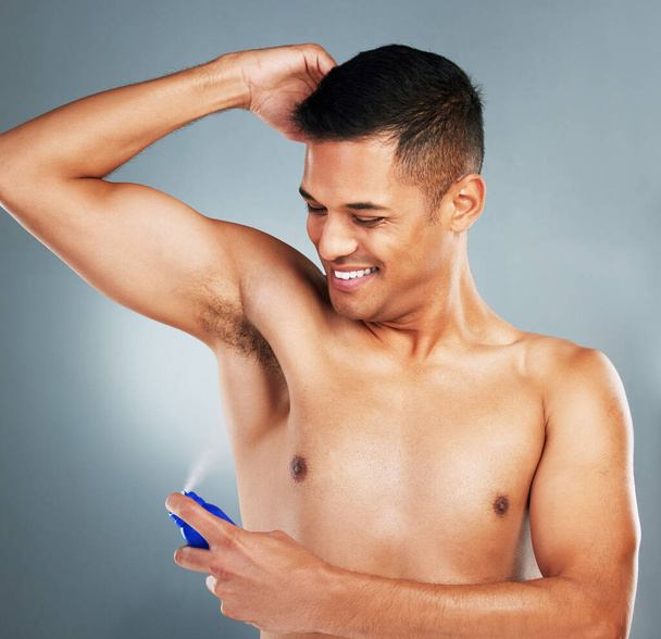 Happy, smile and man with deodorant spray for hygiene, fresh scent and fragrance after a shower. Happiness, clean and guy spraying his armpit to prevent odor or smell isolated by a gray background - Photo, Image