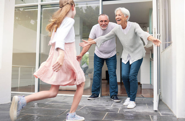 Grandparents, child and welcome for girl running to hug her grandma and grandpa at front door of home. Excited kid bonding, love and hugging senior man and woman for a family visit in Germany. - Foto, Bild
