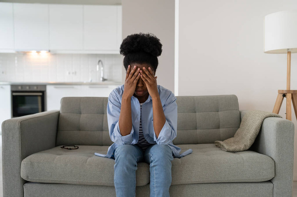 Femme afro-américaine déprimée souffrant de stress et les yeux fermés avec les mains assises sur le canapé dans l'appartement. Jeune fille avec des problèmes psychologiques a besoin de l'aide d'un psychologue après avoir perdu son emploi - Photo, image