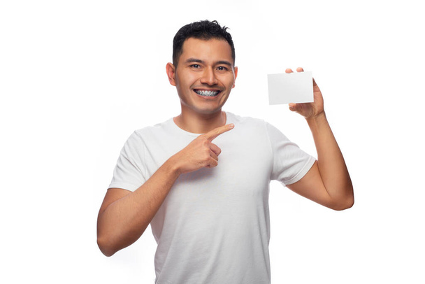 Hombre latino de piel hispana con tratamiento dental de ortodoncia apunta a una tarjeta blanca sobre un fondo claro, una persona - Foto, Imagen