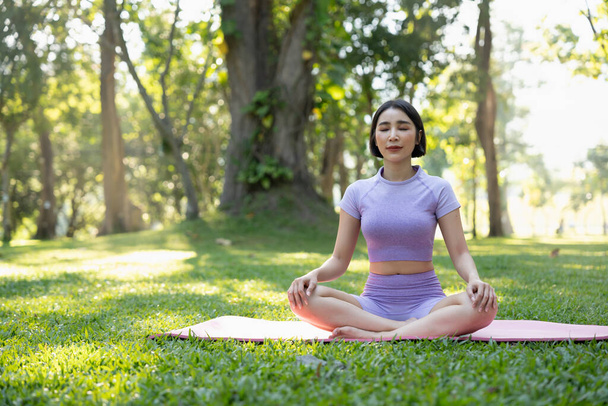 Attractive young Asian woman practice yoga, exercise in the park, standing one leg on a yoga mat, showing balance posture. Wellbeing lifestyle and activity concept. - Photo, Image