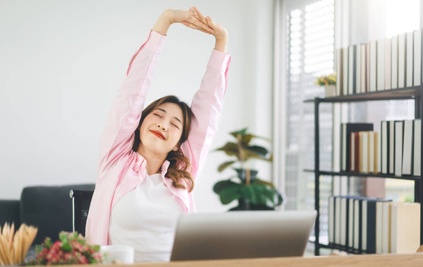 Retrato de jovem adulto sudeste negócio asiático mulher esticando a mão para relaxar o tempo de pausa no local de trabalho. Vestindo camisa rosa casual sentado em casa escritório. - Foto, Imagem
