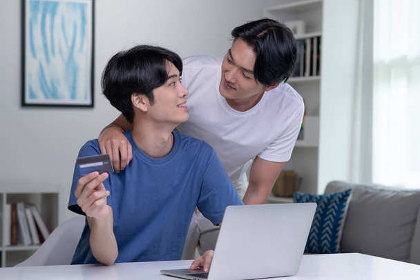 Joven pareja gay asiática sosteniendo tarjeta de crédito y usando computadora portátil. Hombre de negocios gay LGBT trabajando en casa. Compras en línea, comercio electrónico, banca por Internet, gasto de dinero, trabajo desde el concepto de casa - Foto, Imagen