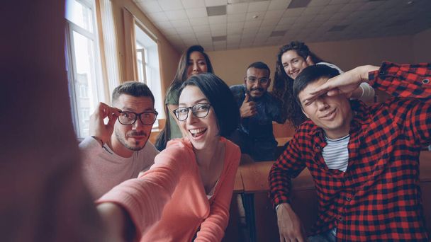 Point of view shot of funny attractive young men and women taking selfie in lecture hall, posing with textbook and showing hand gestures and expressive faces. - Photo, Image