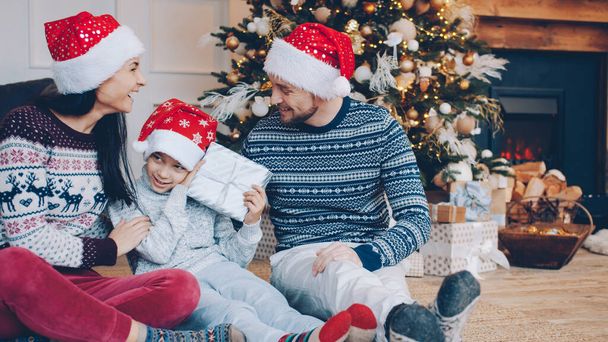 Loving parents in New Year hats are giving present to happy little boy on Christmas eve at home, child is holding decorated box enjoying holiday - Foto, imagen