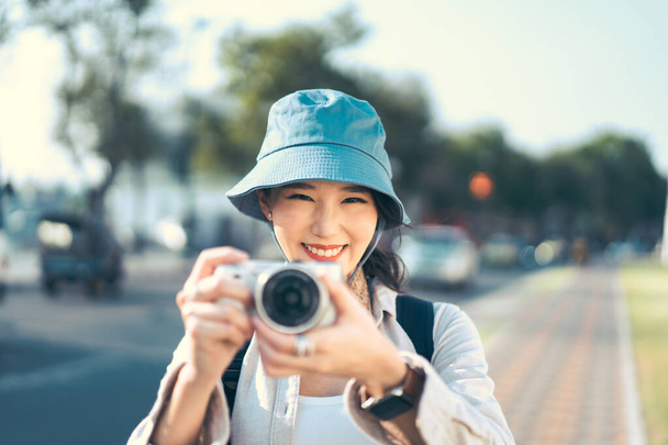 Attrayant sourire heureux jeune voyageur asiatique adulte porter chapeau bleu et sac à dos. Utilisation de l'appareil photo pour le voyage photo. Les gens voyageant dans le mode de vie de la ville le jour. Séjour concept voyage d'été. - Photo, image