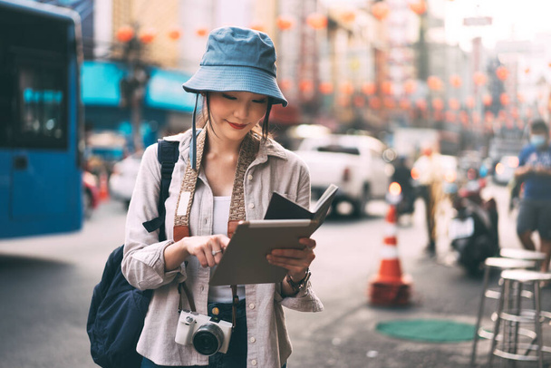 Attractive happy young adult asian woman traveller wear blue hat and backpack. Solo traveling with modern teachnology using tablet for searching lifestyle at city outdoor on day. Summer trip concept. - Zdjęcie, obraz