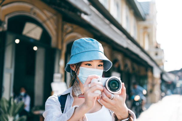 若い大人のアジアの女性のバックパック旅行者は、コロナウイルスの顔マスクや写真のためのカメラを使用してcovid-19女性の人々を着用してください。新しい都市生活の概念. - 写真・画像