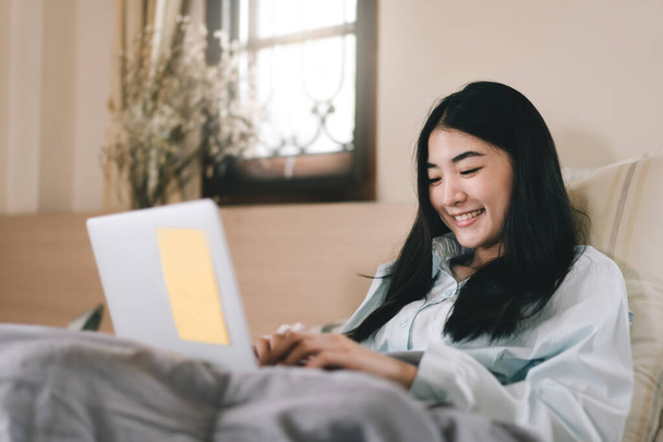Happy smile young adult asian woman using laptop computer for social message and work online. Relax lying on bed in the morning. - Photo, Image
