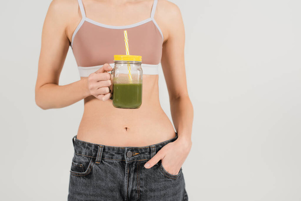 cropped view of young and fit woman holding smoothie while standing with hand in pocket isolated on grey - Photo, Image