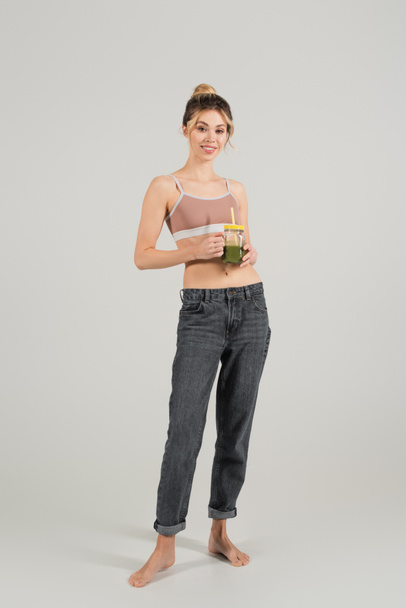 full length of happy barefoot woman in sports top and jeans standing with jar of smoothie on grey background - Фото, изображение