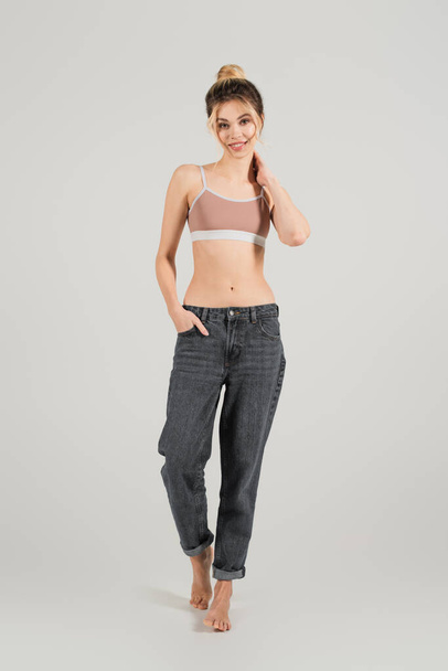 full length of slim barefoot woman in sports top standing with hand in pocket of jeans   on grey background - Photo, Image