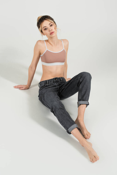 young woman with perfect body and skin sitting in jeans and sports top on grey background - Photo, Image