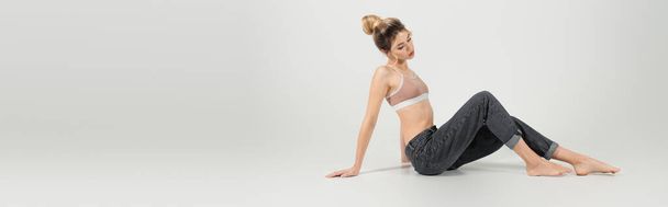 young barefoot woman in jeans and sports top posing on grey background, banner - Фото, изображение