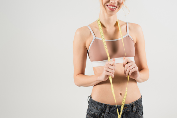 обрезанный вид счастливой и здоровой женщины в спортивном топе проведение измерительной ленты изолированы на серый - Фото, изображение