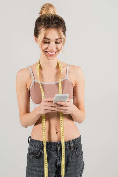 mulher alegre com figura fina e fita métrica usando telefone celular isolado em cinza - Foto, Imagem