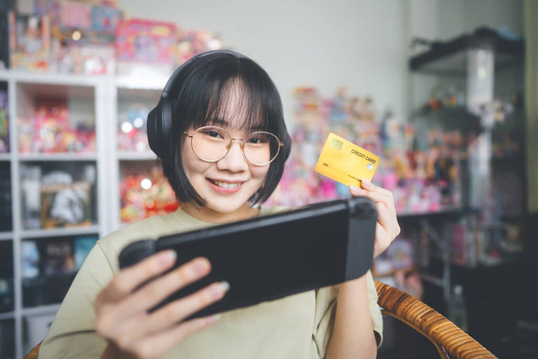 İnek stili genç Asyalı oyuncu kadın gözlük takıyor ve kulaklık takıyor. Online bir oyun oynuyor. Yarışı kazanmak için kredi kartını kullanmak.. - Fotoğraf, Görsel