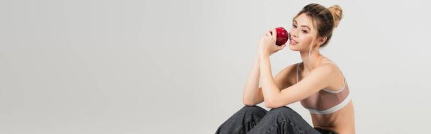 pozytywna młoda kobieta w bluzce sportowej siedzi z dojrzałym jabłkiem odizolowanym na szarym, sztandarze - Zdjęcie, obraz