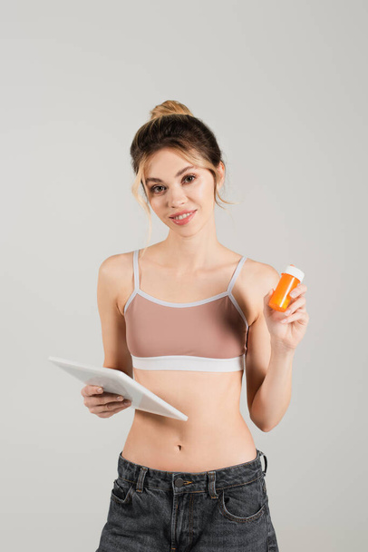 en forma y saludable mujer en sujetador deportivo celebración tableta digital y vitaminas aisladas en gris - Foto, Imagen
