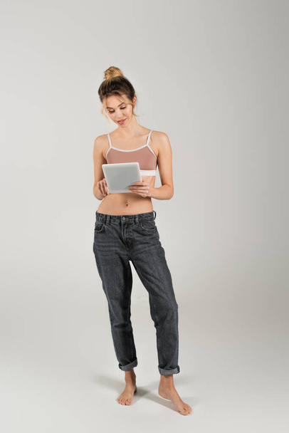 full length of fit barefoot woman in jeans and sports top using digital tablet on grey background - Фото, зображення