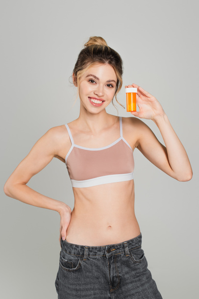 cheerful and fit woman in sports top standing with hand on hip and showing container with vitamins isolated on grey - Photo, Image