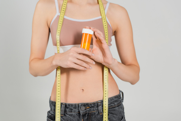 partial view of slim woman with measuring tape and container with vitamins isolated on grey - Foto, imagen