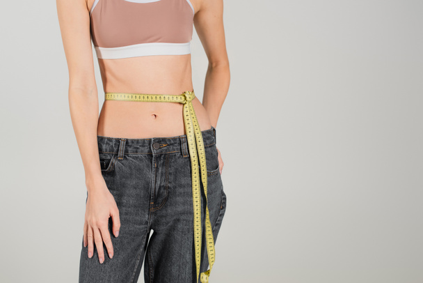 partial view of fit woman in sports top and jeans standing with measuring tape on waist isolated on grey - Photo, Image