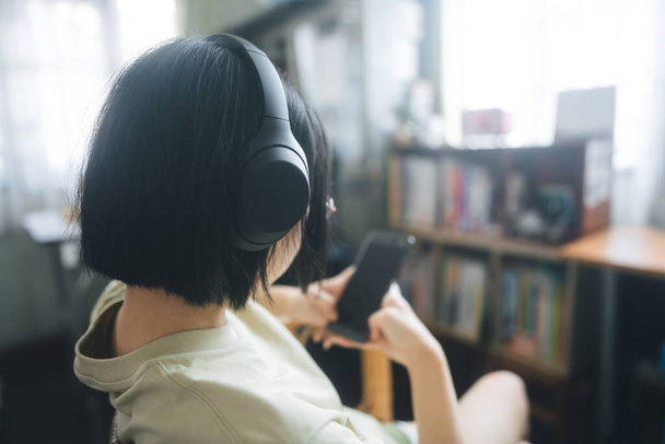 Οι άνθρωποι ευτυχισμένη τρόπο ζωής ικανοποίηση στο σπίτι έννοια. Νεαρή ενήλικη Ασιάτισσα γυναίκα φορούν ακουστικά ακούσετε μουσική και να χαλαρώσετε χρησιμοποιώντας ψηφιακό κινητό τηλέφωνο για σερφάρισμα στο διαδίκτυο και online ψώνια. - Φωτογραφία, εικόνα