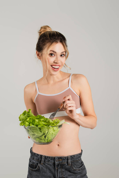 cheerful woman in sports top holding bowl of lettuce and fork isolated on grey - Fotoğraf, Görsel