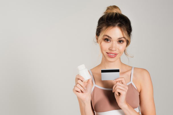 Doğal makyajlı ve mükemmel ciltli güzel bir kadın kredi kartı ve vitaminleri gri renkte tutuyor. - Fotoğraf, Görsel