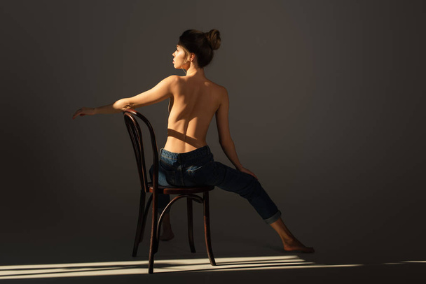 back view of half nude barefoot woman in jeans posing on wooden chair on grey background with lighting - Photo, Image