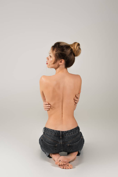 back view of half naked barefoot woman in jeans sitting and looking away on grey background - Photo, Image