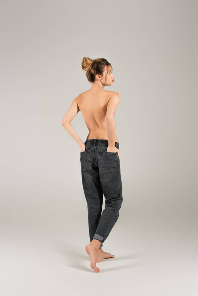vista trasera de mujer descalza medio desnuda de pie con las manos en los bolsillos traseros de jeans sobre fondo gris - Foto, Imagen