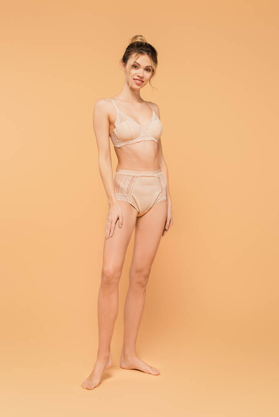full length of fit woman in lace underwear smiling at camera on beige background - Photo, Image