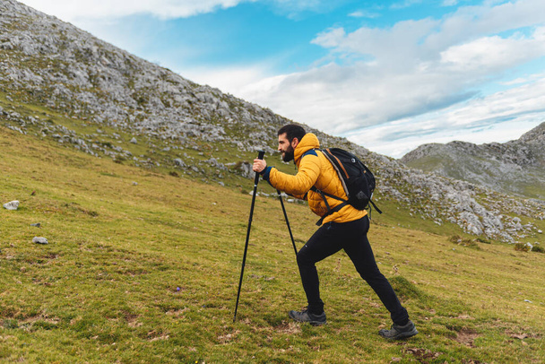Caucasian man with backpack and yellow jacket, hiking with trekking poles. mountaineer ascending a mountain peak. healthy lifestyle - Photo, Image