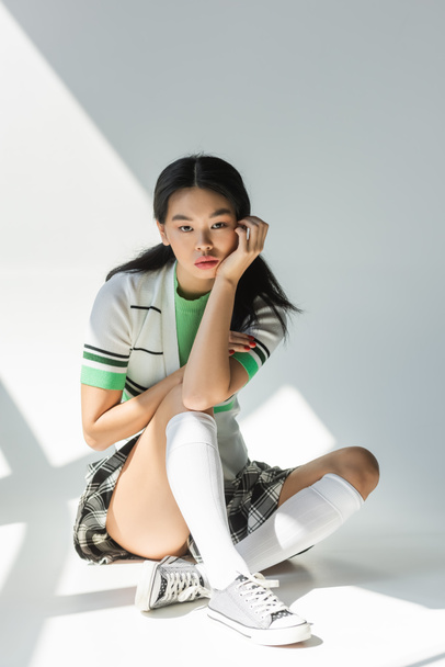 Asian woman in skirt and knee socks sitting on grey background with light  - Foto, Imagem
