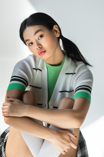 Молодая азиатка в полосатом кардигане обнимает колени на сером фоне  - Фото, изображение