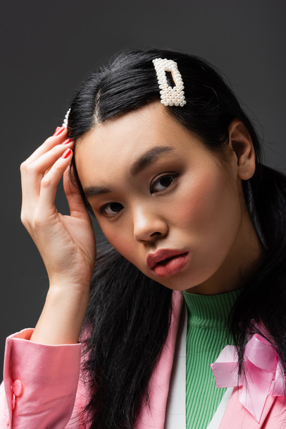 Portrait of asian model adjusting barrette and looking at camera isolated on grey  - Photo, Image